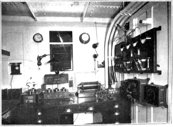 A ship radio room with conduits for wires an a wooden office desk. On the desk and on the walls are wooden boards and boxes with knobs and switches